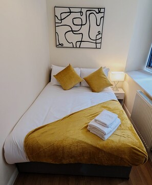 Un lit bien fait, recouvert d’une couvre-lit blanche et jaune moutarde, avec deux coussins jaunes moutarde et une serviette blanche pliée posée sur le lit.