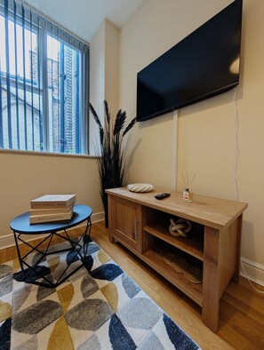Ein Wohnzimmer mit einem Wand-Fernseher, einem Holzschrank mit Regalen, einem kleinen runden Tisch und einem blauen Stuhl sowie einem gemusterten Teppich.