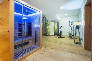 Ein Saunaraum mit Glastrennwand, Holzboden und Trainingsgeräten im angrenzenden Bereich.