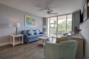Ein Wohnzimmer mit blauem Sofa, einem Couchtisch, einem Beistelltisch, einem Deckenventilator und einem Balkon mit Gartenmöbeln.