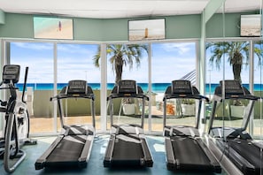 A fitness area with treadmills and exercise bikes, overlooking a beach with palm trees and a clear sky. - Steps to the Sand! Gulf Front Studio at Boardwalk Resort – Pool, Hot Tubs, Tiki Bar & More! (Panama City Beach)
