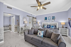 A spacious living area with a grey sofa, a ceiling fan, and a bed with nautical-themed decor. - Steps to the Sand! Gulf Front Studio at Boardwalk Resort – Pool, Hot Tubs, Tiki Bar & More! (Panama City Beach)