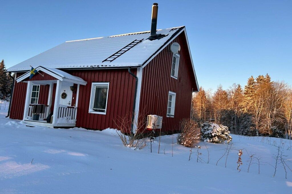 A red wooden house with a snow-covered roof, a chimney, and a small porch with a door and windows.