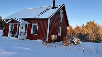 A red wooden house with a snow-covered roof, a chimney, and a small porch with a door and windows.