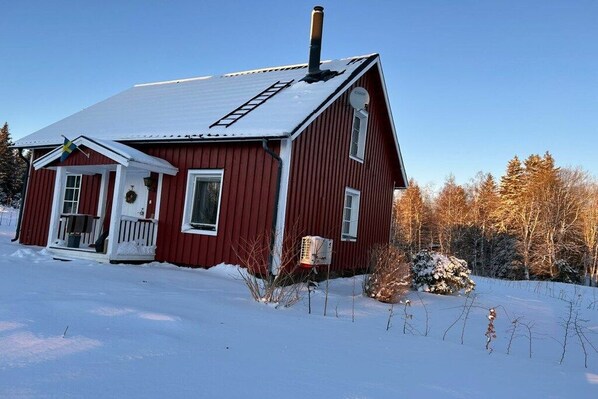 A red wooden house with a snow-covered roof, a chimney, and a small porch with a door and windows.