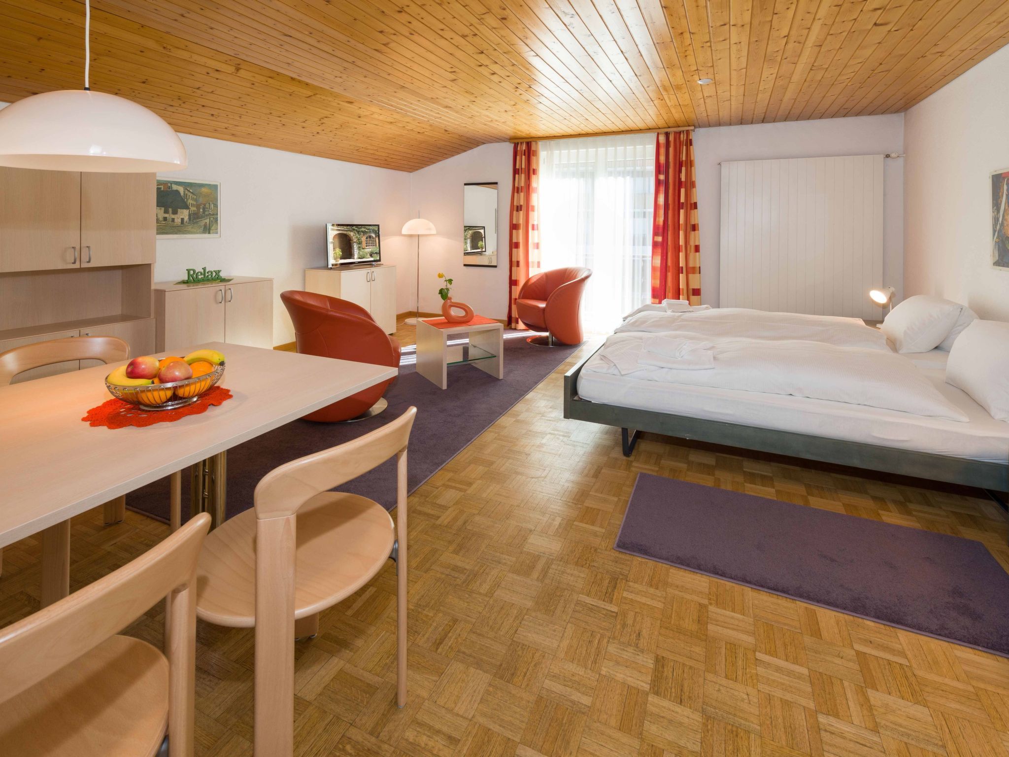 Quarto de hotel moderno com teto de madeira, área de refeições com mesa e cadeiras, cama e televisão.