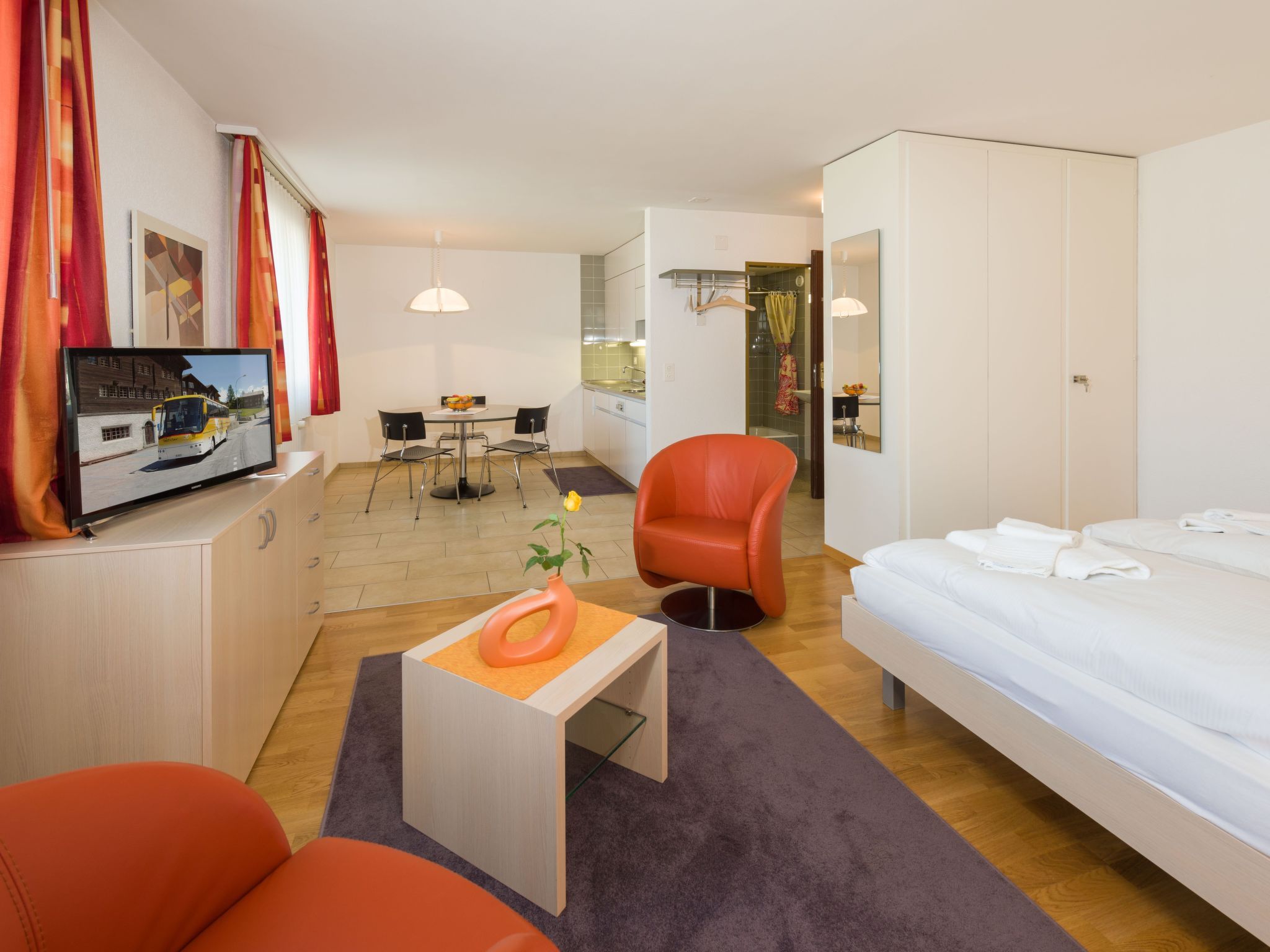 A hotel room with a bed, a desk, a chair, a television, and a kitchen area.