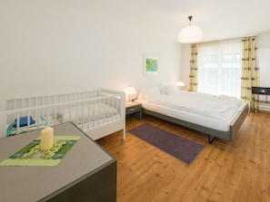 Une chambre à coucher avec un berceau, un lit, un bureau avec une lampe et une fenêtre avec des rideaux.