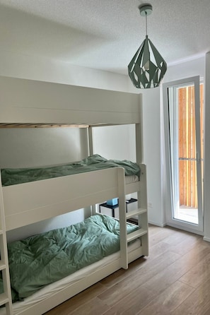 Ein Etagenbett mit einer grünen Matratze, ein kleiner Tisch und eine Hängeleuchte in einem Zimmer mit Holzboden und einem fenster mit Vorhängen.