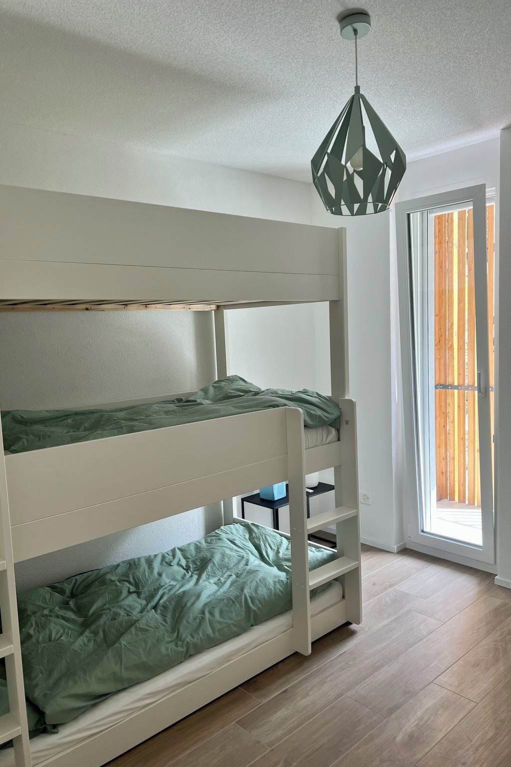 Una cama individual superpuesta con escritorio plegable, lámpara colgante y puerta que da a un balcón.