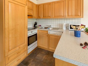 A kitchen with wooden cabinets, a stove, a dishwasher, and a countertop with a coffee maker and utensils. - Försterli West, (Grächen), 3002 by Interhome (Grächen)
