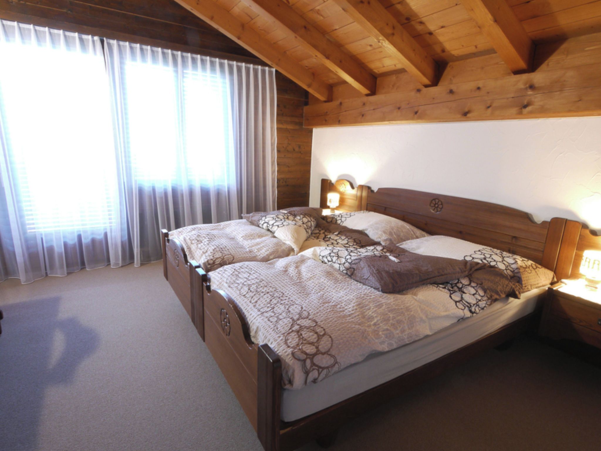 Ein Schlafzimmer mit Holzbettrahmen, einem großen Bett, zwei Nachttischlampen und einem Fenster mit durchsichtigen Vorhängen.