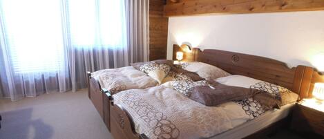 Ein Schlafzimmer mit Holzbettrahmen, einem großen Bett, zwei Nachttischlampen und einem Fenster mit durchsichtigen Vorhängen.