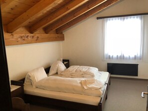 A bedroom with a wooden ceiling, a bed with white bedding, a nightstand, and a window with sheer curtains. - Ferienwohnungen Gsteinetgufer, (Grächen), by Interhome (Grächen)