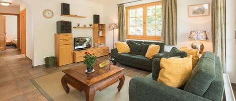 Ein Wohnzimmer mit einer Couch, einem Couchtisch und einem Fernseher. Ein Fenster mit Vorhängen, eine Uhr an der Wand und eine Tür zu einem anderen Raum.