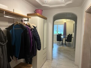 Un couloir mène à une salle à manger où se trouvent une table et des chaises, un porte-manteau avec différents vestons, et une étagère en bois sur laquelle est posé un seau rose.