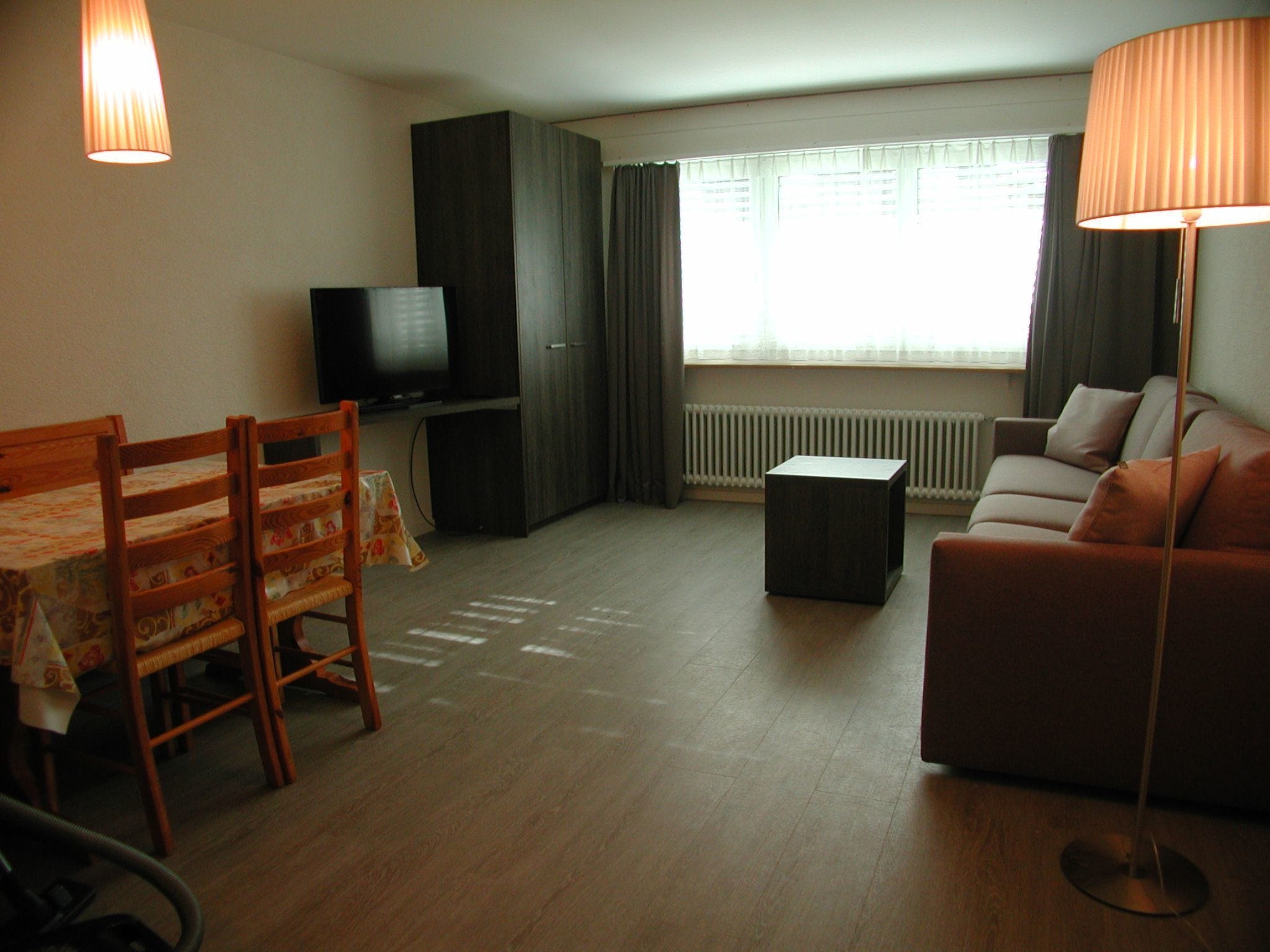 A furnished living room with a sofa, a television, a dining table with chairs, and a lamp.