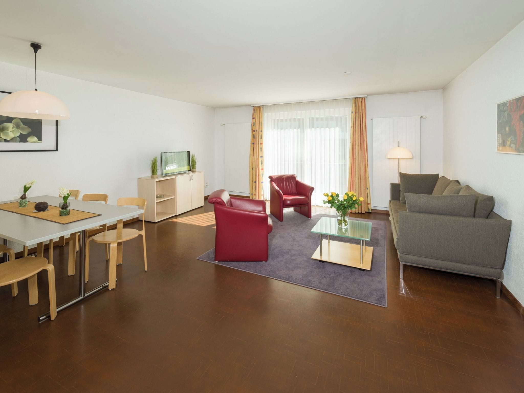 Una sala moderna con comedor, un sofá de cuero rojo, un sofá gris, una mesa de centro de vidrio y un televisor sobre un mueble de madera.