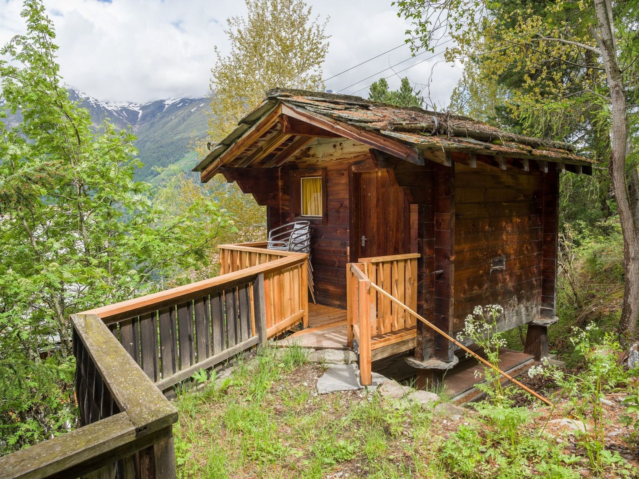 A wooden cabin with a wooden deck and a single window, surrounded by trees and mountains.