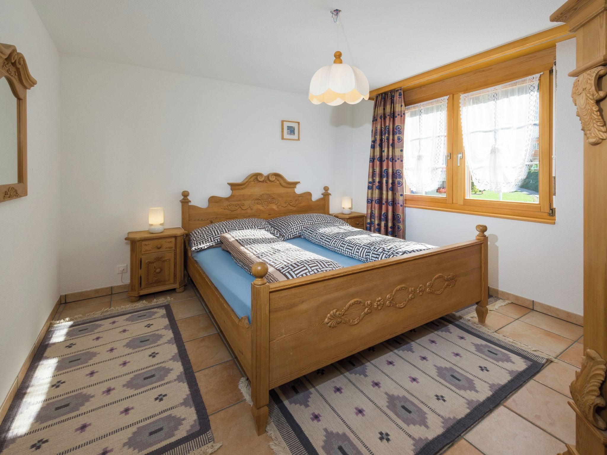 A bedroom with a wooden bed, bedside lamps, a patterned rug, and a window with curtains.