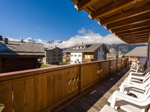 A wooden balcony with white plastic chairs, overlooking a mountain village with traditional buildings and a snow-capped peak in the background. - Paradiso 2, (Grächen), 2003 by Interhome (Grächen)