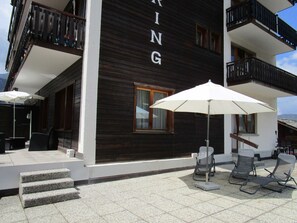 A building with a dark wooden facade, a balcony with a black railing, and outdoor seating with umbrellas. - Touring, (Grächen), 1030 by Interhome (Grächen)