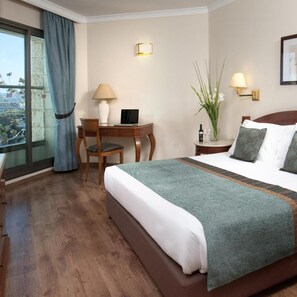 A hotel room with a bed, a desk, a chair, a window with curtains, and a nightstand with a lamp. - Holiday Inn (Haifa)