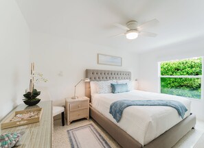 Une chambre à coucher comprenant un lit, des tables de chevet, une petite table, une lampe sur pied, un ventilateur de plafond, une fenêtre donnant sur un espace verdoyant et une petite plante.