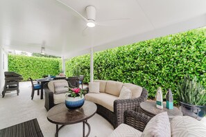 An outdoor patio area with a ceiling fan, a round table, a sofa, and a chair. There is a green hedge wall in the background. - New Boutique Juno Retreat | Pool I Steps to Beach! (Juno Beach)
