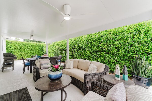 An outdoor patio area with a ceiling fan, a round table, a sofa, and a chair. There is a green hedge wall in the background. - New Boutique Juno Retreat | Pool I Steps to Beach! (Juno Beach)
