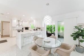 A modern kitchen with white cabinetry, a central island, and a dining area with a glass table and chairs. - Sea Turtle Sanctuary by the Beach! Heated Pool! (Juno Beach)