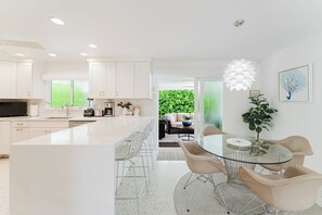 A modern kitchen with white cabinetry, a central island, and a dining area with a glass table and metal chairs. - Sea Turtle Sanctuary by the Beach! Heated Pool! (Juno Beach)
