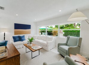 A modern living room with a white sofa, a grey armchair, and a wooden coffee table. There is a large window with a view of greenery outside. - New Boutique Juno Retreat | Pool I Steps to Beach! (Juno Beach)