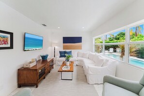 Un salon moderne avec un canapé blanc, une table basse en bois et une grande fenêtre donnant sur une piscine et un espace verdoyant.