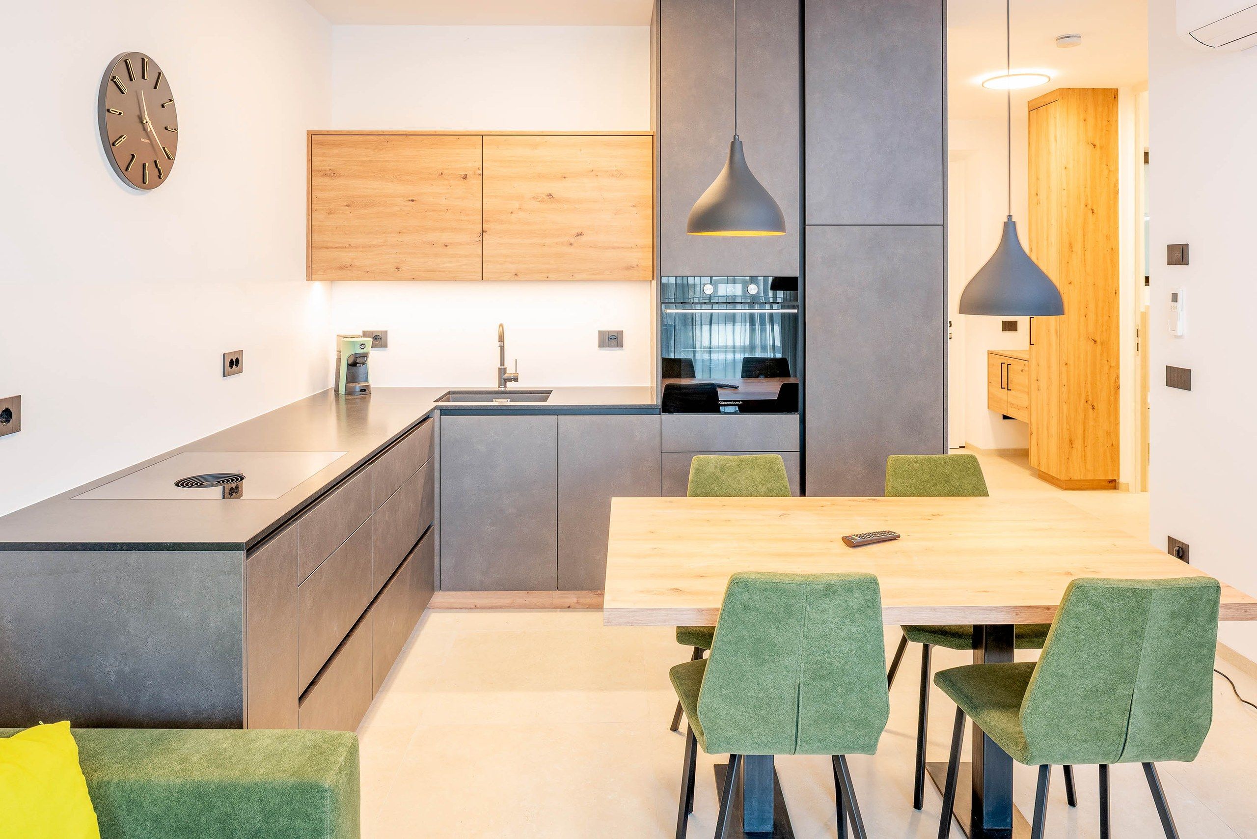 Une cuisine moderne avec un îlot central, un coin repas avec une table en bois et des chaises à revêtement vert, et une horloge au mur.