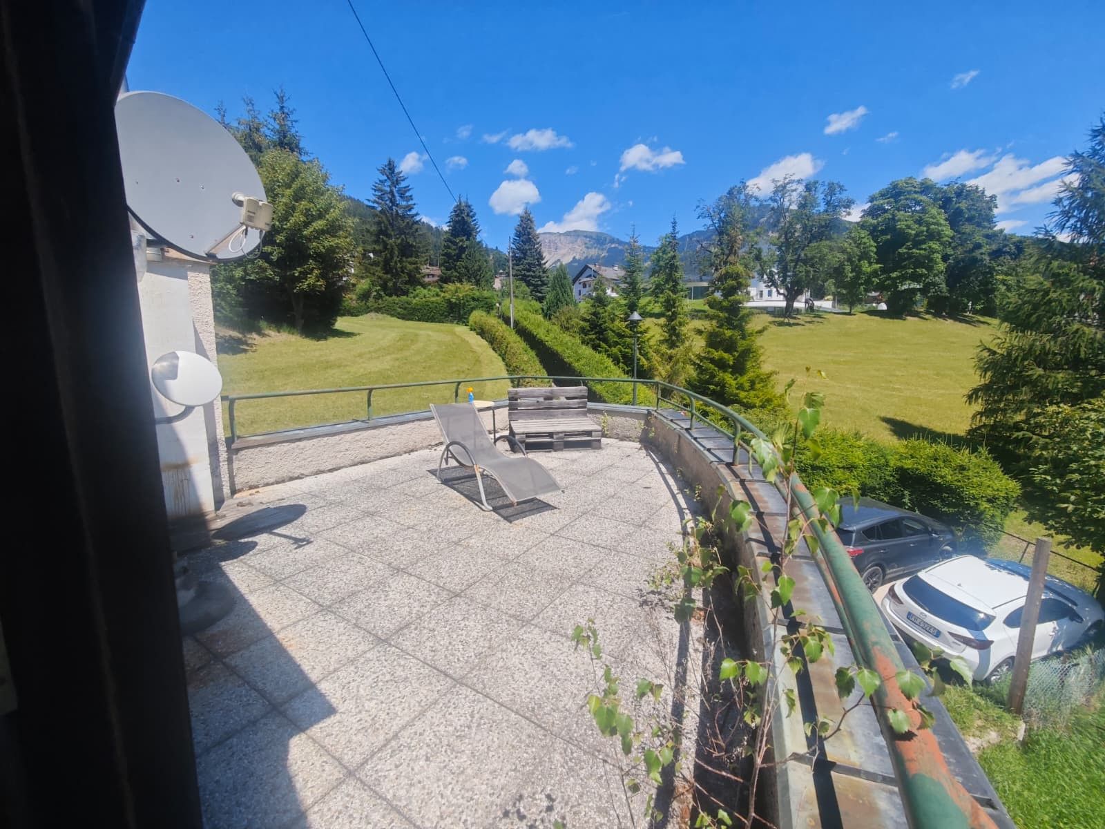Un balcon donnant sur un paysage verdoyant, une antenne parabolique et une voiture garée à l’extérieur.
