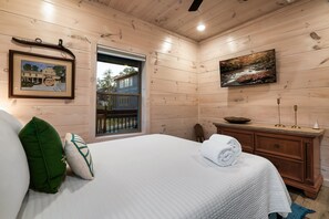 Ein Zimmer mit Holzwänden, einem Bett, einem Gemälde, einem Fenster und einer Holzkommode.