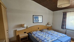 A bedroom with a wooden headboard, two bedside tables with lamps, a bed with blue bedding, a framed picture on the wall, and a window with curtains.