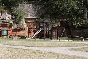 Un terrain de jeux avec un toboggan et des structures d’escalade, le tout adossé à un mur de pierre et entouré d’arbres.