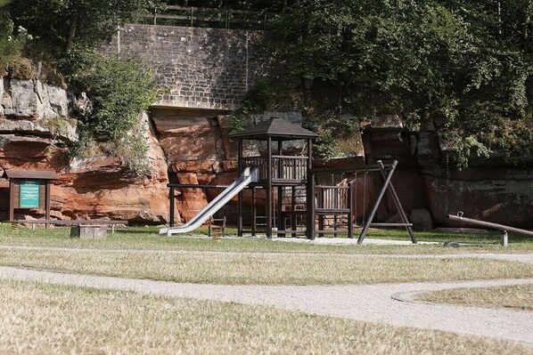 A playground with a slide and climbing structures, set against a backdrop of a stone wall and trees.