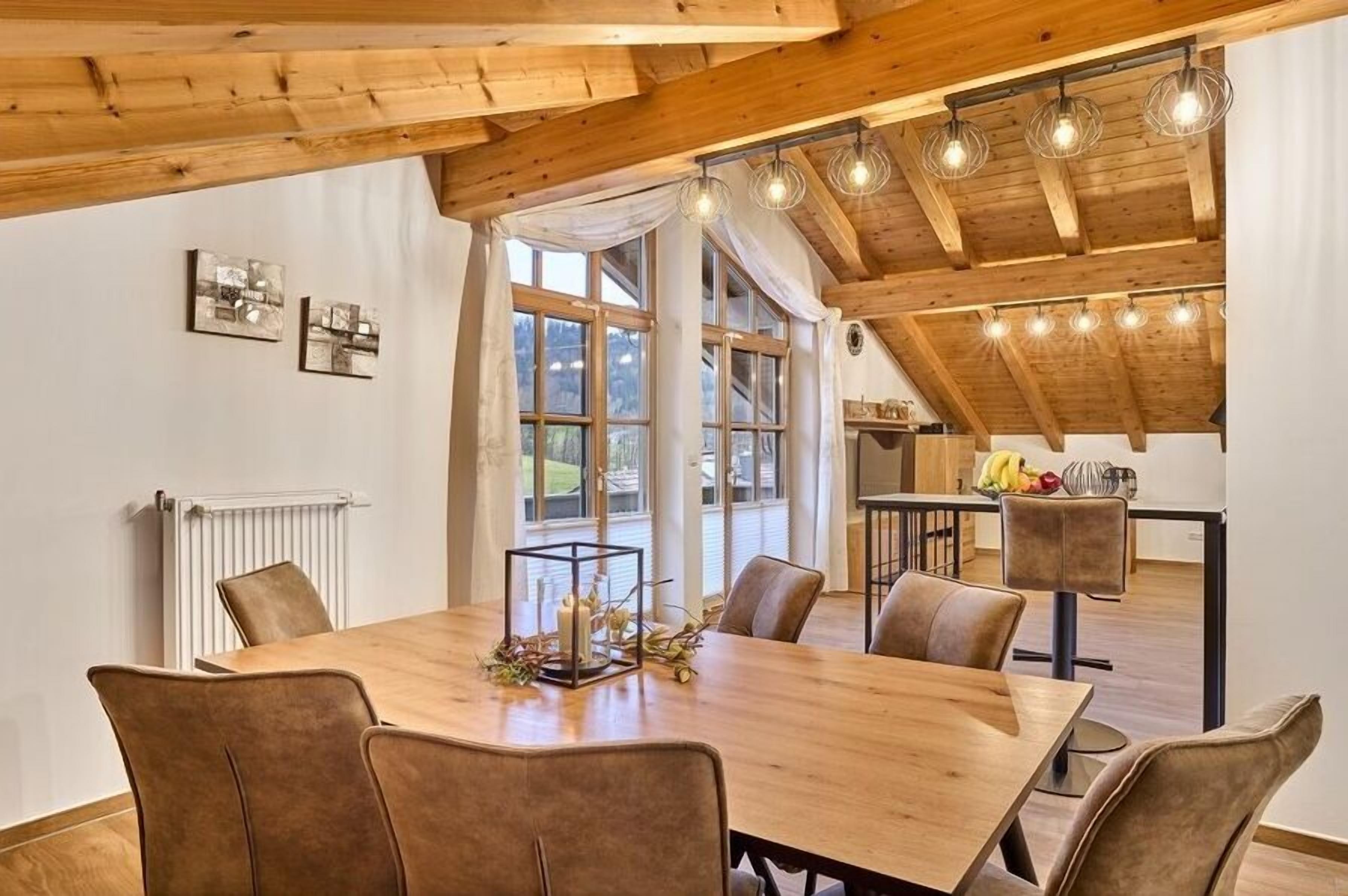 A dining area with a wooden table and chairs, a kitchen in the background, and large windows.