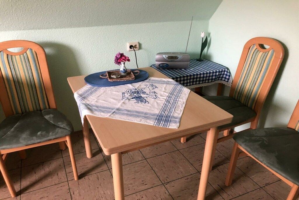 Una zona de menjador amb una taula preparada per a dues persones, dues cadires, una taula petita amb un mantell a quadres i un aparell de ràdio penjat a la paret.