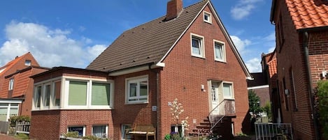 A red brick residential building with a sloped roof, a front porch, and a small garden area.