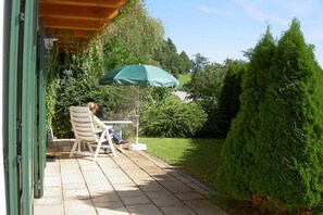 Pihalla on patio, jossa on pöytä ja tuoli, vihreä aurinkovarjo sekä hyvin hoidettu puutarha, jonka keskellä kohoaa suuri havupuu.