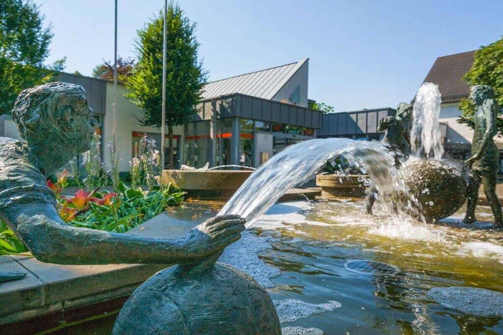 Une statue représentant une figure tenant un récipient d’eau, une fontaine, et un bâtiment en arrière-plan.