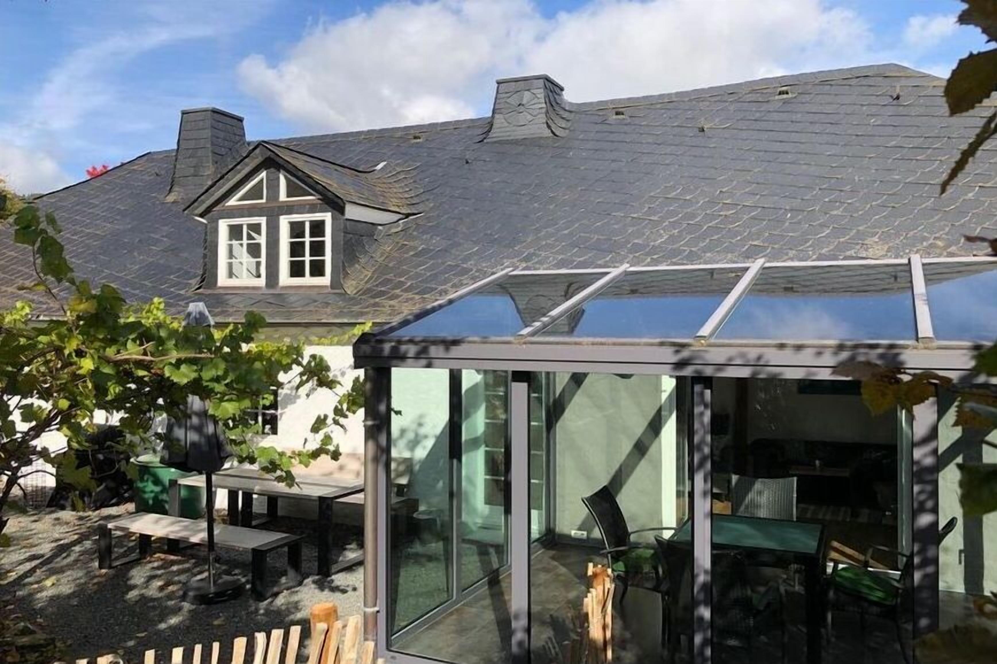 Ein Wintergarten mit Glasdach, ein Essbereich mit Gartenmöbeln und im Hintergrund ein Haus mit Satteldach.