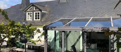 Ein Wintergarten mit Glasdach, ein Essbereich mit Gartenmöbeln und im Hintergrund ein Haus mit Satteldach.