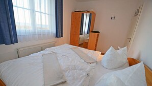 A hotel room with a bed, a wooden wardrobe, a window with curtains, and a radiator. - Right on the Baltic Sea beach with a view of the sea!, 20 meters to the beach, washing machine, sleeps 2 (Glowe)