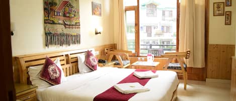 A hotel room with a large bed, a wooden headboard, a painting on the wall, a window with curtains, and a view of buildings outside.