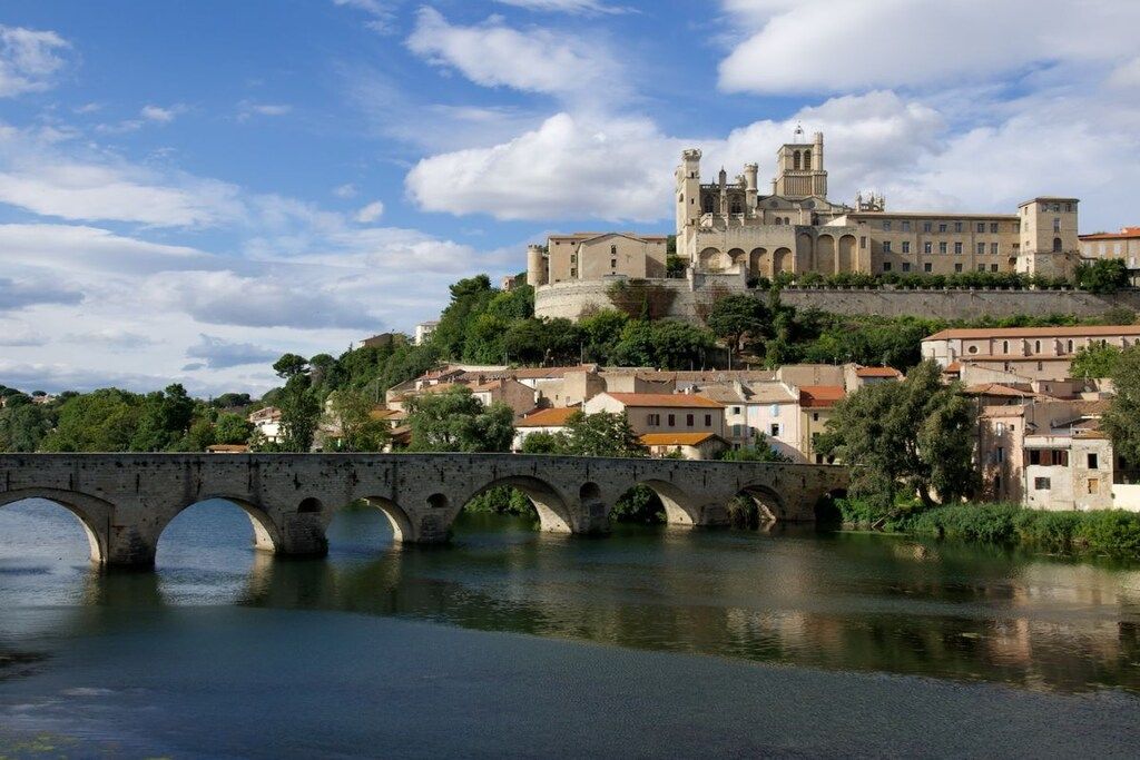 A historic bridge with multiple arches spanning a river, leading to a fortified castle with towers and a large building with multiple windows.